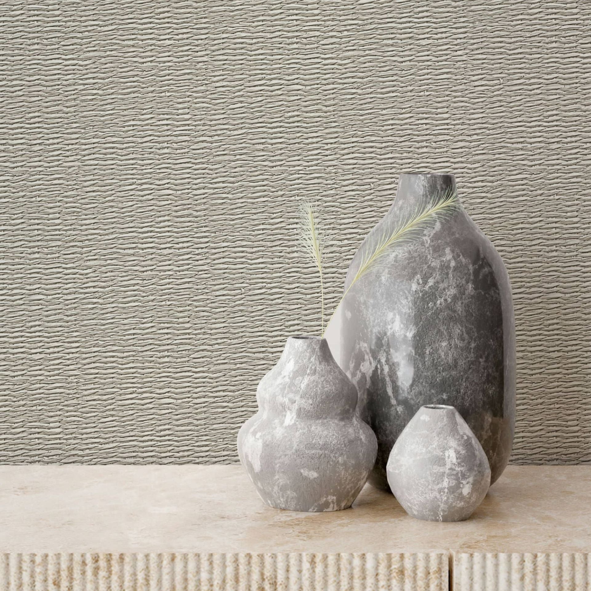 Marburg Travertino 33383 Textured beige wallpaper with narrow horizontal ridges behind three gray stone vases on a light stone surface.