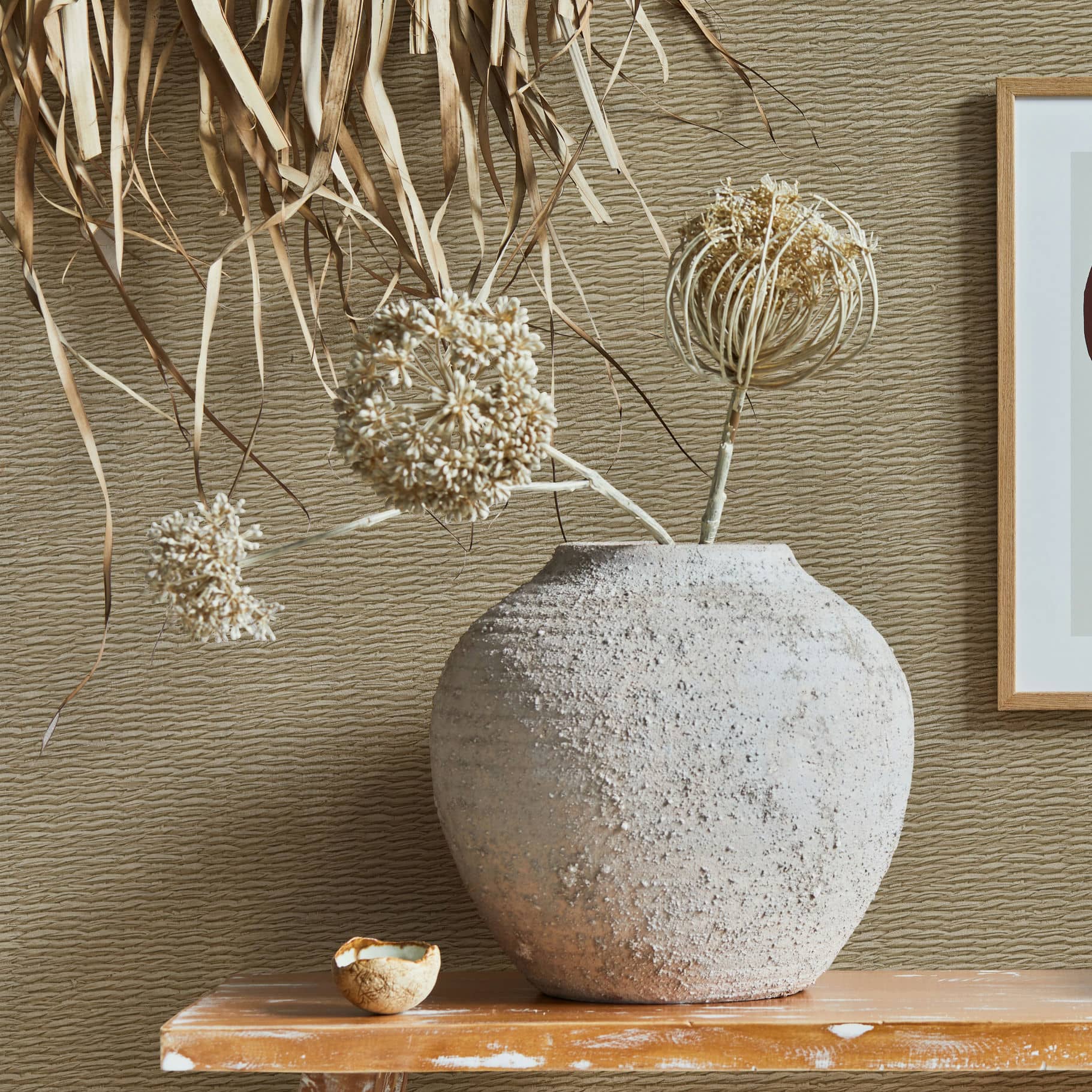 Marburg Travertino 33384 Beige wallpaper with fine horizontal ripples shown behind a large rustic ceramic vase with dried stems on a wooden shelf, a small bowl, hanging dried leaves, and the edge of a framed print.