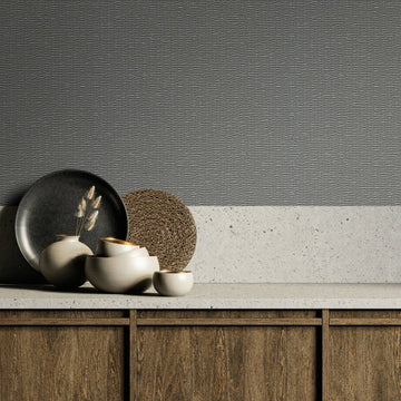 Marburg Travertino 33386 Gray textured wallpaper with horizontal ripple lines shown above a light stone countertop and backsplash, with ceramic bowls, a small vase with dried stems, a dark plate, a woven placemat, and wood cabinets below.