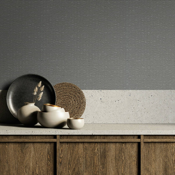 Marburg Travertino 33386 Gray textured wallpaper with horizontal ripple lines shown above a light stone countertop and backsplash, with ceramic bowls, a small vase with dried stems, a dark plate, a woven placemat, and wood cabinets below.
