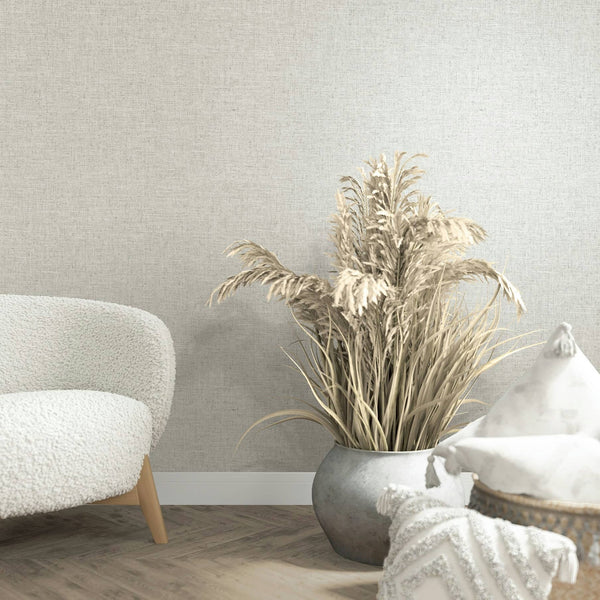 Marburg Travertino 33388 Light beige-gray linen-look textured wallpaper behind a boucle chair, a floor planter of dried grasses, and decorative pillows on a wood floor with baseboard trim.