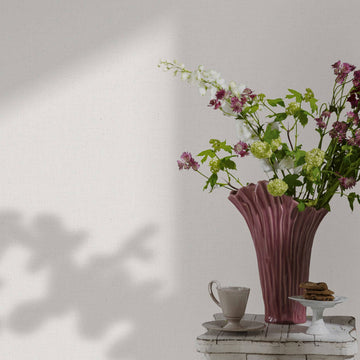 Marburg Travertino 33390 Light white textured wallpaper with a soft fabric-like weave, shown behind a pink sculptural vase of flowers on a small table with a teacup and cookies. Leaf shadows fall across the wall.