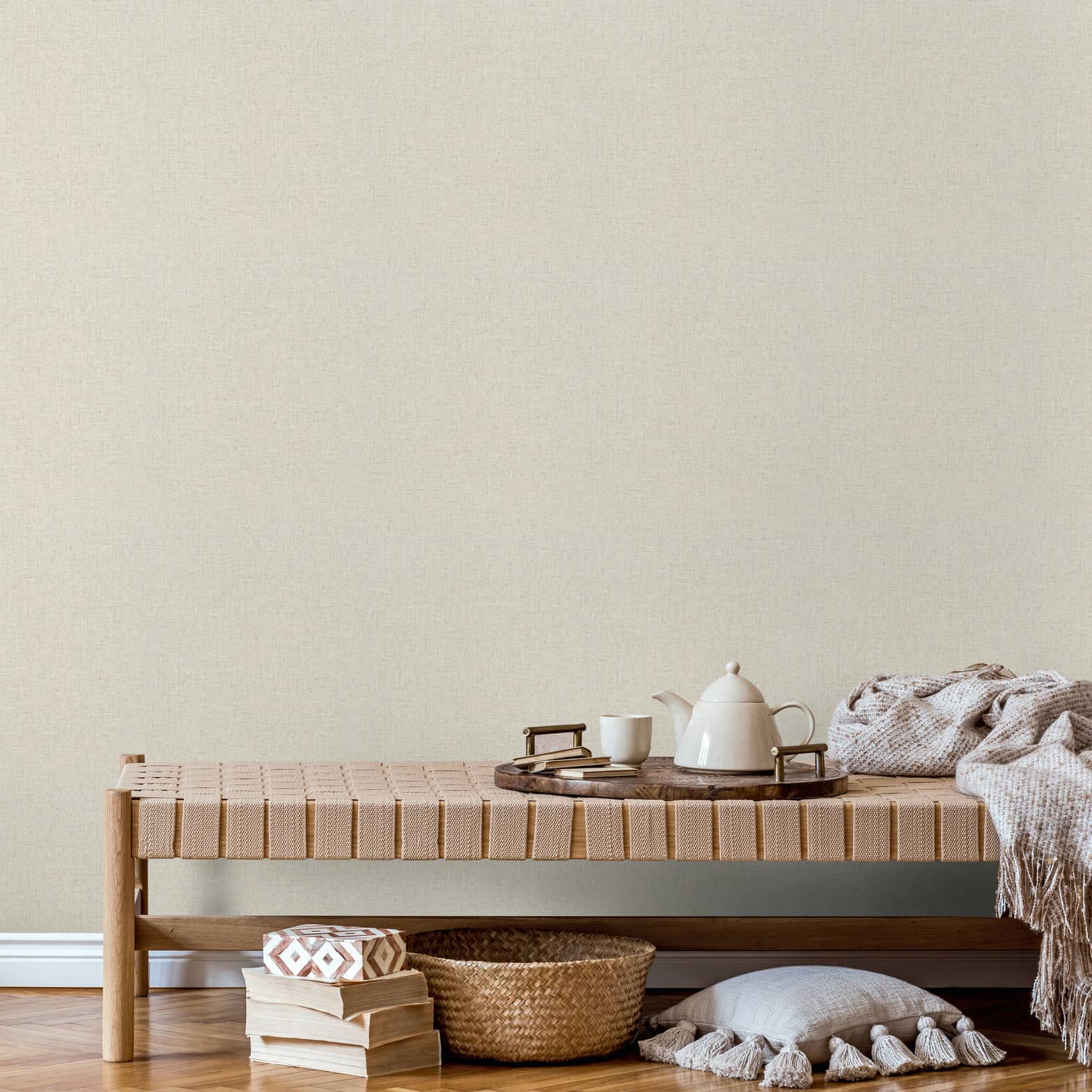 Marburg Travertino 33391 Light beige textured wallpaper behind a woven bench styled with a tray, teapot, and cup, with a throw, basket, stacked books, and a tasseled floor cushion by a wall with a white baseboard.