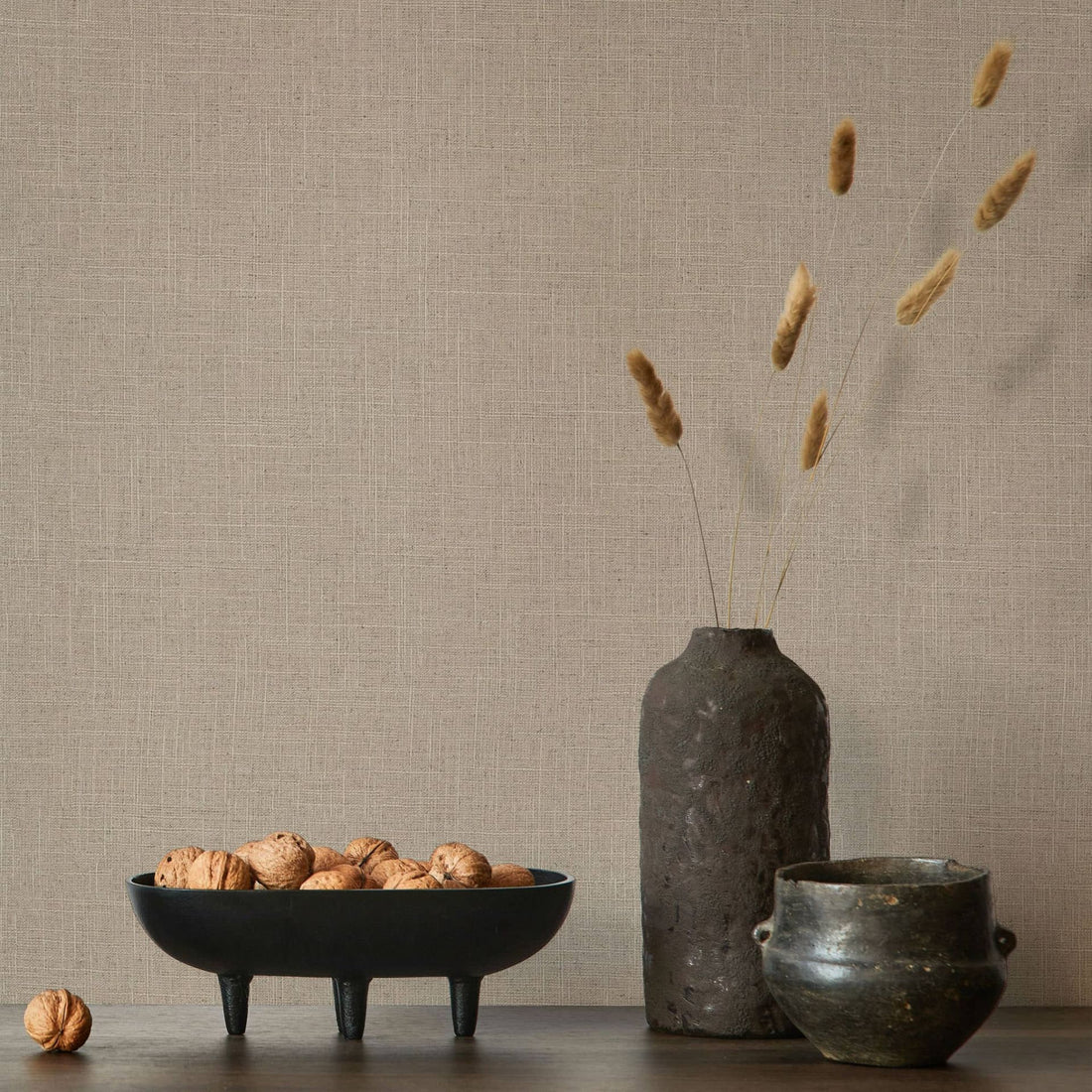 Marburg Travertino 33393 Warm brown crosshatch-textured wallpaper behind a tabletop scene with a black footed bowl of walnuts, a tall dark vase with dried stems, and a small dark pot.
