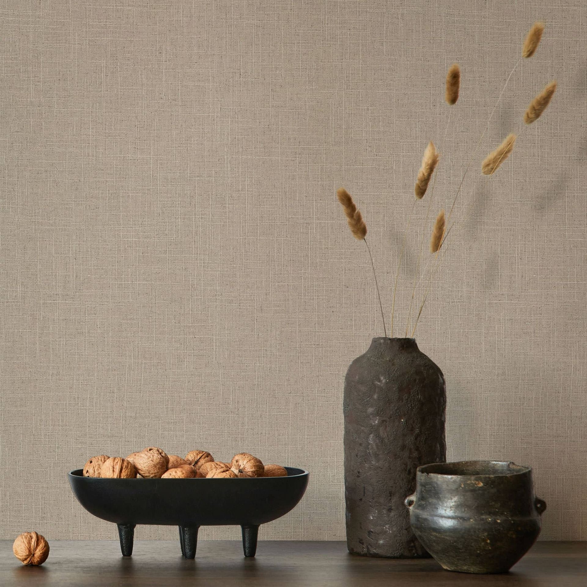 Marburg Travertino 33393 Warm brown crosshatch-textured wallpaper behind a tabletop scene with a black footed bowl of walnuts, a tall dark vase with dried stems, and a small dark pot.
