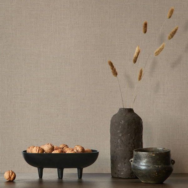 Marburg Travertino 33393 Warm brown crosshatch-textured wallpaper behind a tabletop scene with a black footed bowl of walnuts, a tall dark vase with dried stems, and a small dark pot.