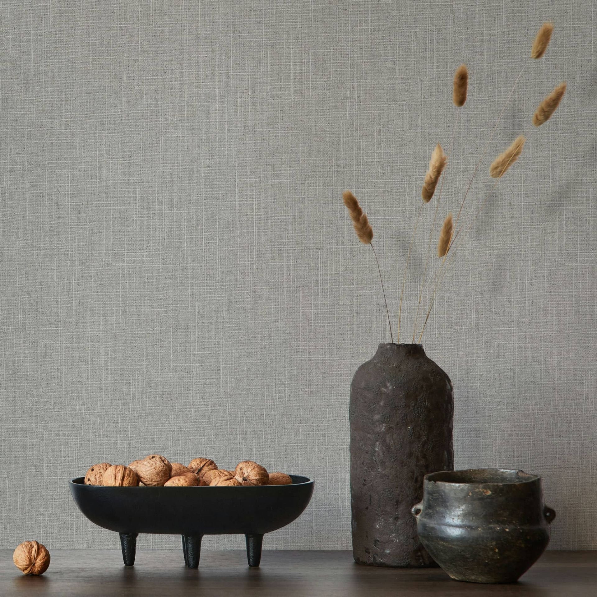 Marburg Travertino 33394 Gray crosshatch wallpaper behind a dark ceramic vase with dried grasses, a small pot, and a black bowl filled with walnuts on a wooden surface.