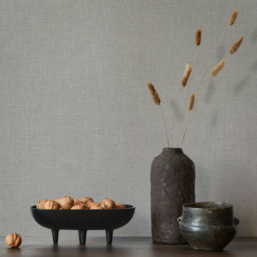 Marburg Travertino 33394 Gray crosshatch wallpaper behind a dark ceramic vase with dried grasses, a small pot, and a black bowl filled with walnuts on a wooden surface.