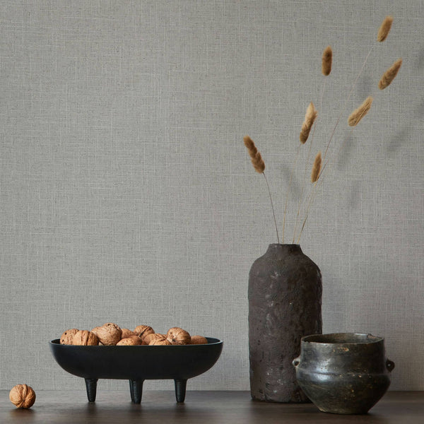 Marburg Travertino 33394 Gray crosshatch wallpaper behind a dark ceramic vase with dried grasses, a small pot, and a black bowl filled with walnuts on a wooden surface.