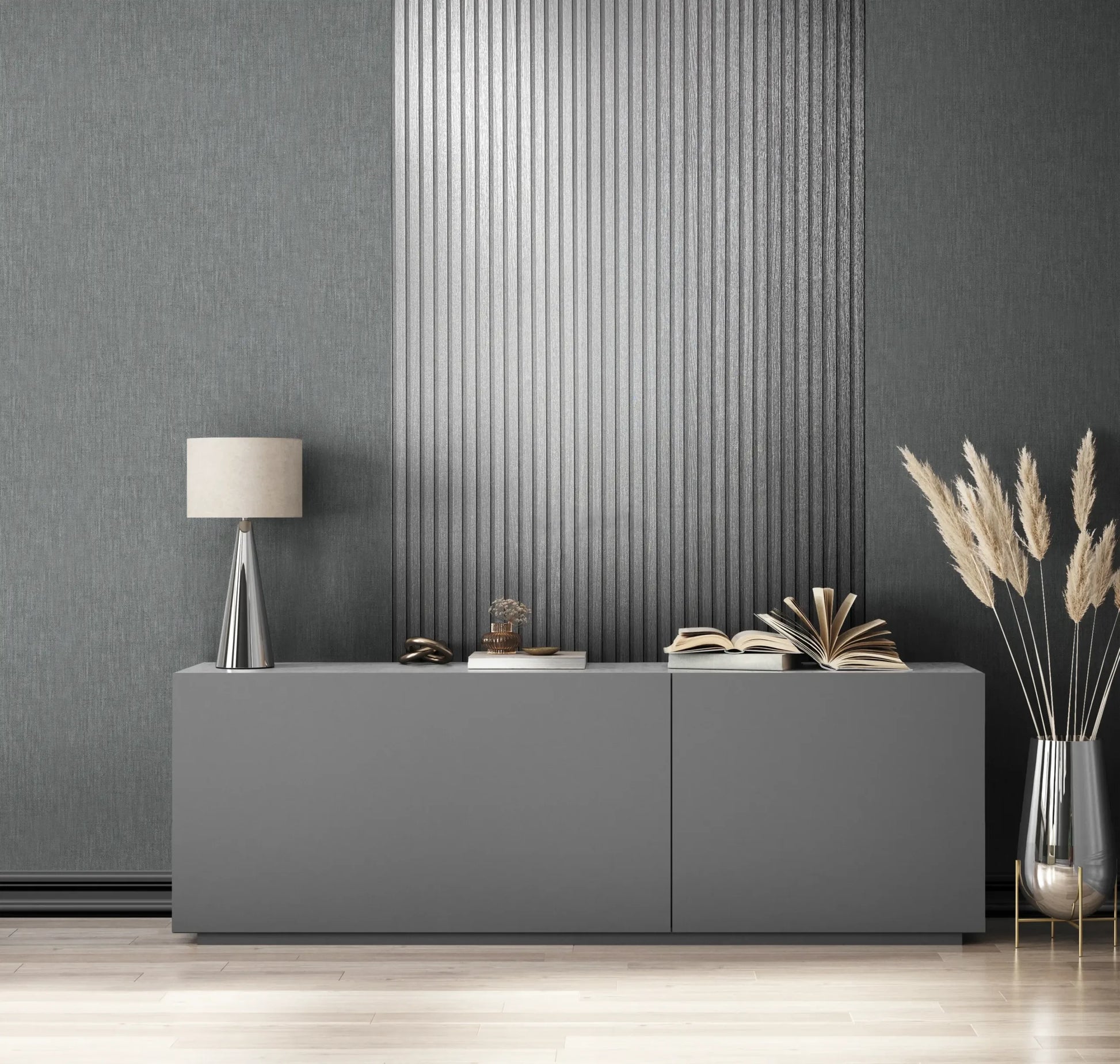 Marburg Travertino 33395 Room scene with textured dark grey wallpaper and a central vertical panel of metallic silver ribbing behind a low grey sideboard, a cone-based table lamp, stacked and open books, and a tall glass floor vase with pampas stems.