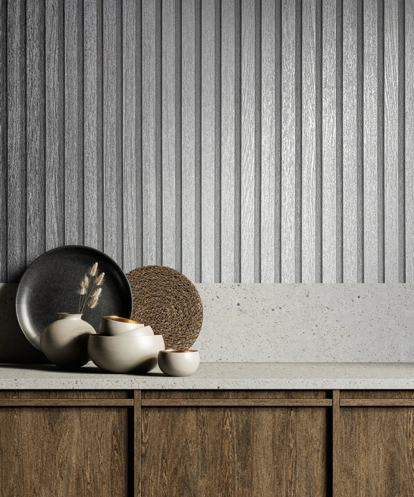 Marburg Travertino 33395 Textured metallic silver vertical‑rib wallpaper behind a light stone countertop with wood cabinets, ceramic vessels, a dark plate, and a round woven mat.