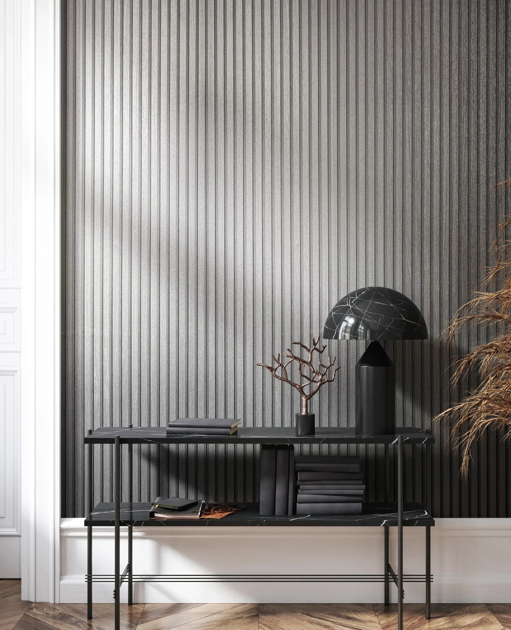 Marburg Travertino 33395 Vertical ribbed silver wallpaper behind a black console table with a black dome lamp, stacked books, a small decorative branch, and dried foliage; white trim and a herringbone wood floor are visible.