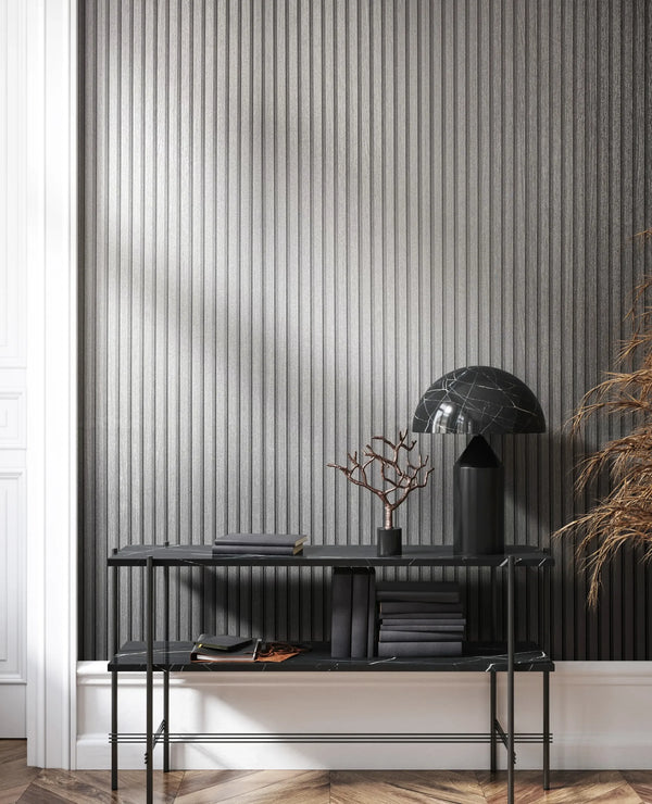 Marburg Travertino 33395 Vertical ribbed silver wallpaper behind a black console table with a black dome lamp, stacked books, a small decorative branch, and dried foliage; white trim and a herringbone wood floor are visible.
