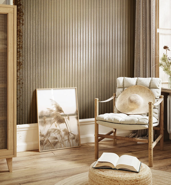 Marburg Travertino 33396 Gold-toned vertical ribbed wallpaper shown as a feature wall behind a cushioned wooden chair with a hat, a leaning framed print, a window with curtains, and a woven pouf with an open book.