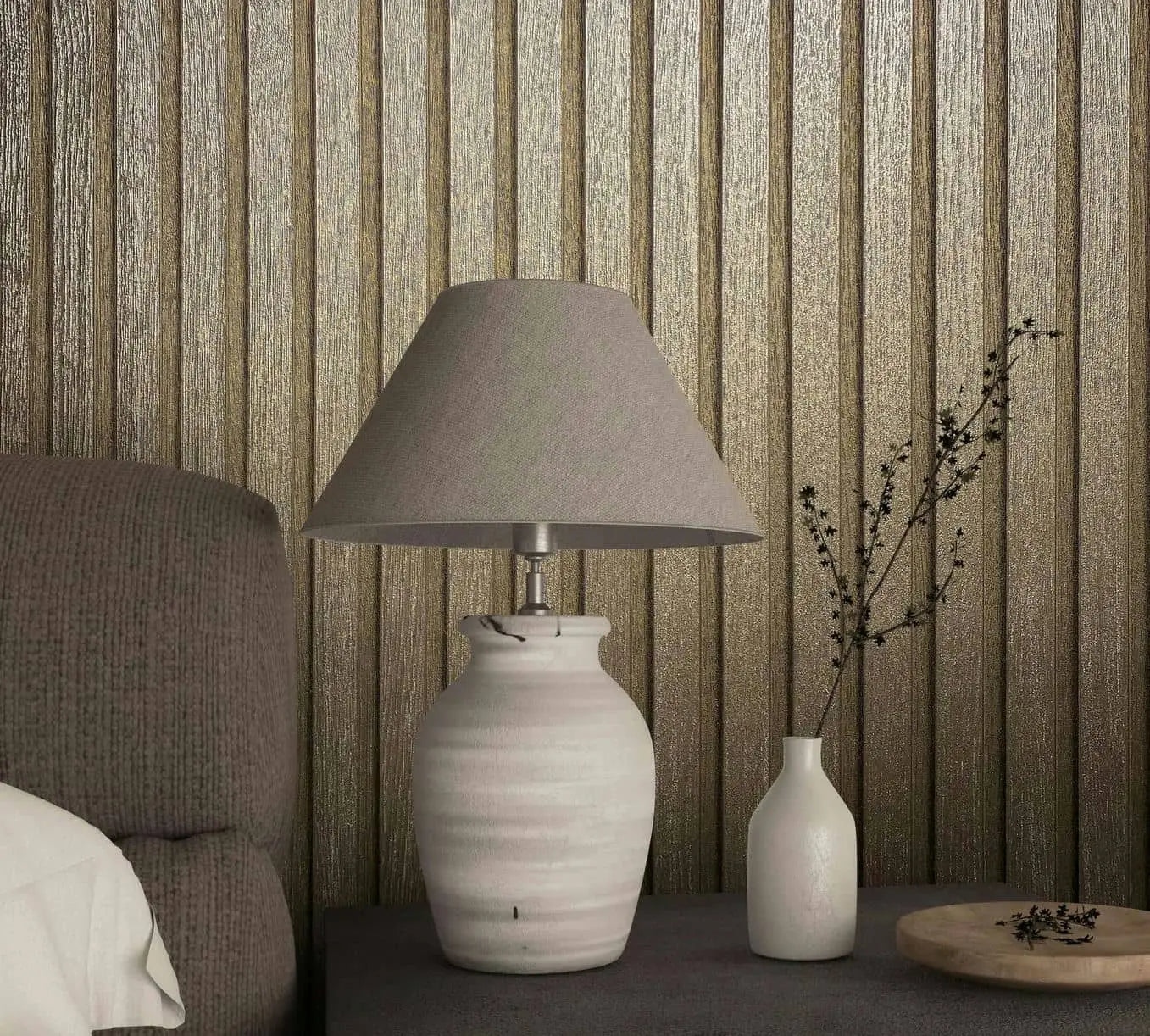 Marburg Travertino 33396 Metallic gold vertical ribbed wallpaper behind a side table with a ceramic lamp, a small white vase holding a branch, a round wooden tray, and the edge of a textured sofa.