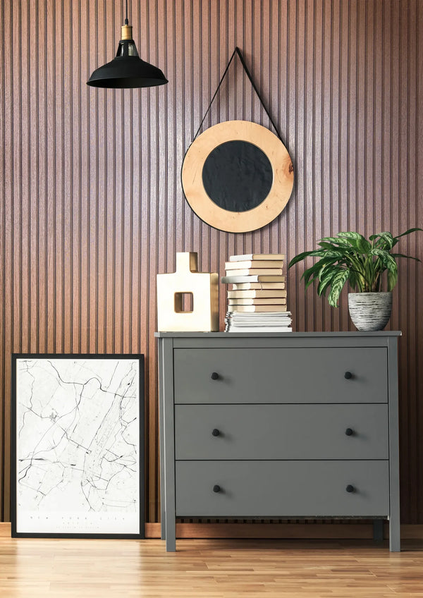 Marburg Travertino 33397 Vertical ribbed copper-toned wallpaper shown in a styled room with a gray three-drawer dresser, round mirror on a strap, black pendant light, stacked books, a decorative object, a potted plant, and a framed map print on the floor.