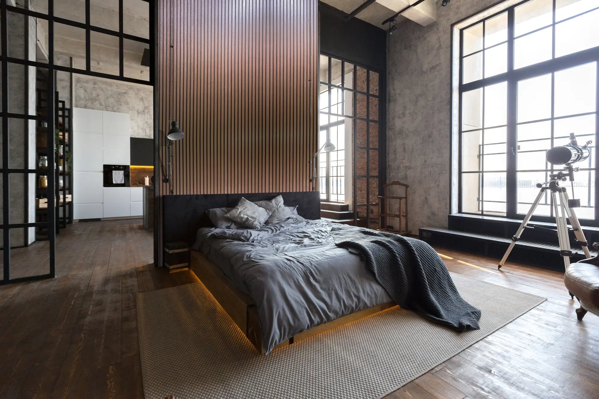 Marburg Travertino 33397 Industrial loft bedroom with a bed set against a panel of copper-toned vertical stripe wallpaper, large grid windows, and a telescope on a tripod by the window.